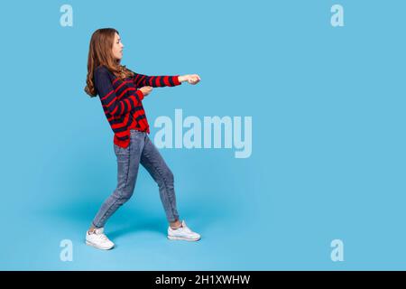 Portrait complet de femme portant un chandail rayé de style décontracté, debout dans l'attaque ou tirant les mains gestuelle et regardant vers l'avant.Studio d'intérieur isolé sur fond bleu. Banque D'Images