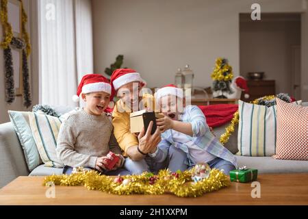 Un père caucasien et deux fils ouvrent une boîte cadeau pendant un appel vidéo sur tablette numérique pendant noël Banque D'Images