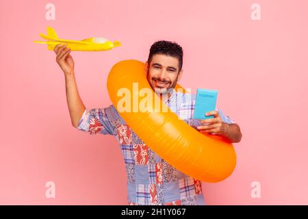 heureux touriste satisfait en chemise bleue avec anneau en caoutchouc, tenant un passeport et une maquette d'avion, profitant de la visite de voyage.Studio d'intérieur isolé sur fond rose. Banque D'Images