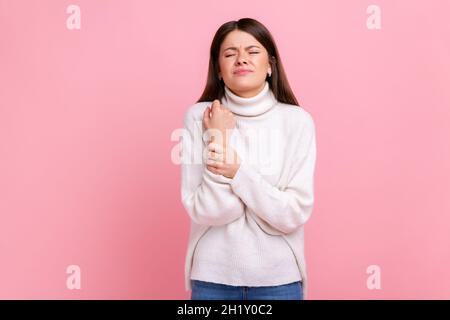 Femme contrariée debout avec la grimace de douleur, massant mal de poignet, souffrant de blessure de main ou de crasse, portant blanc style décontracté pull.Studio d'intérieur isolé sur fond rose. Banque D'Images