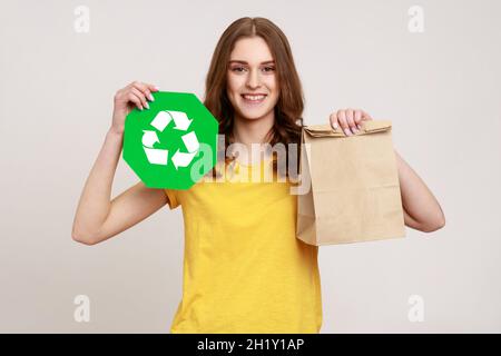 Jeune fille souriante portant un T-shirt jaune décontracté tenant le paquet de papier et l'affiche verte de recyclage, regardant l'appareil photo, exprimant le bonheur.Prise de vue en studio isolée sur fond gris. Banque D'Images