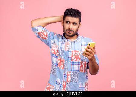 Homme barbu réfléchi en chemise bleue à la tête égratigante tenant le smartphone à la main, en envisageant la mise à jour de logiciels, en choisissant des tarifs appropriés.Studio d'intérieur isolé sur fond rose. Banque D'Images