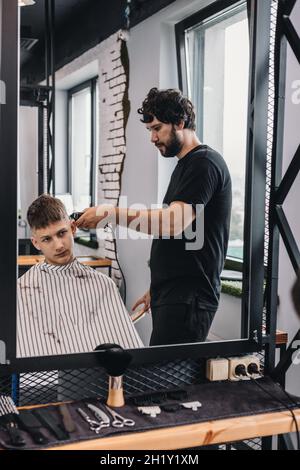 Le coiffeur brunette barbu donne une coupe de cheveux tendance à un adolescent.Réflexion dans le miroir.Coupe de cheveux et coiffage professionnels pour hommes dans un barbe Banque D'Images