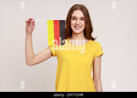 Portrait d'une jeune fille à l'aspect agréable en T-shirt jaune tenant le drapeau de l'Allemagne, célébrant le jour de l'Allemagne - 3 octobre, exprimant positif.Prise de vue en studio isolée sur fond gris. Banque D'Images