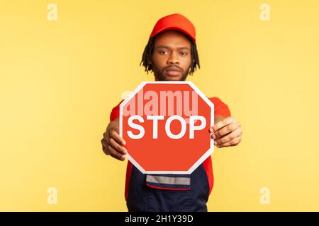 Portrait d'un homme de main sérieux portant un uniforme et un capuchon rouge de visière regardant la caméra, tenant dehors montrant le panneau rouge d'arrêt de circulation, avertit de danger.Studio d'intérieur isolé sur fond jaune. Banque D'Images