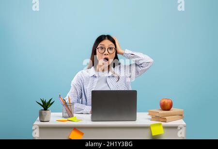 Concept d'échéance.Choqué femme asiatique assise à un bureau avec un ordinateur portable et saisissant sa tête sur fond bleu Banque D'Images