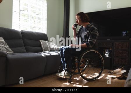 Homme caucasien attentionné assis sur un fauteuil roulant pour boire du café à la maison Banque D'Images