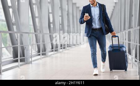Jeune homme d'affaires méconnaissable utilisant un smartphone lors d'une marche avec Suitcase à l'aéroport Banque D'Images