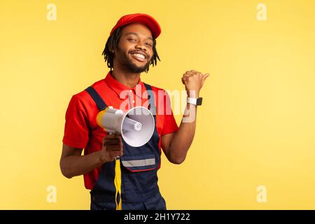 Joyeux homme à barbe portant une combinaison bleue et un T-shirt rouge tenant le mégaphone et pointant le doigt de côté, en regardant sourire l'appareil photo.Studio d'intérieur isolé sur fond jaune. Banque D'Images