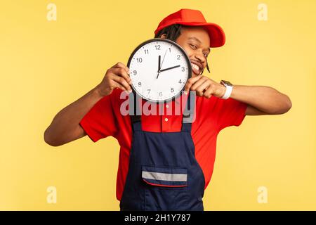 Souriant, un homme de main souriant portant un uniforme bleu tenant une grande horloge murale entre les mains, assure que le travail soit mal fait à temps, industrie du service.Studio d'intérieur isolé sur fond jaune. Banque D'Images