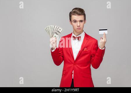 Portrait d'un homme d'affaires fier et beau avec une élégante coiffure dans une veste rouge et un noeud papillon montrant de l'argent et une carte de banque, vous avez l'air confiant et riche. Studio en intérieur tourné isolé sur fond gris Banque D'Images