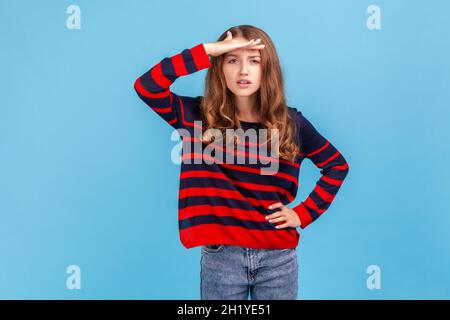 Essayer de voir quelque chose à distance.Femme portant rayé style décontracté chandail tient la paume sur le front regarde loin recherches quelqu'un focalisé loin loin loin.Studio d'intérieur isolé sur fond bleu. Banque D'Images