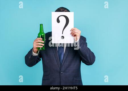 Portrait d'une personne anonyme portant un costume de style officiel foncé se cachant derrière le papier avec un point d'interrogation et tenant une bouteille avec de l'alcool.Studio d'intérieur isolé sur fond bleu. Banque D'Images