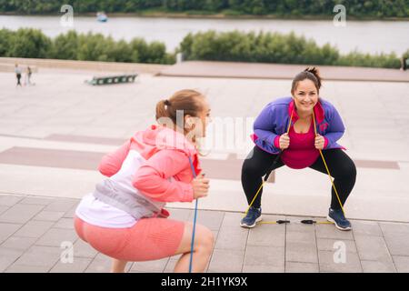 Prise de vue large de la jeune femme en surpoids faisant des exercices de squats à l'aide de ruban de fitness pour la perte de poids avec entraîneur personnel en plein air dans la journée d'été. Banque D'Images