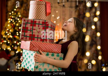 Excitée curieuse petite fille souriante, tenant pile de cadeaux de Noël.Chambre affectueuse joliment décorée avec des lumières et un sapin de Noël.Joyeuses vacances Banque D'Images