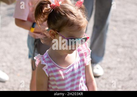 Petite fille triste belle en lunettes de soleil et une robe rayée sur une promenade Banque D'Images