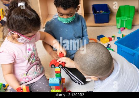 Éducation préscolaire 3-4 ans deux garçons et une fille jouant dans la zone de bloc avec des véhicules jouets et une construction qu'ils ont construit à partir de la connexion colorée b Banque D'Images