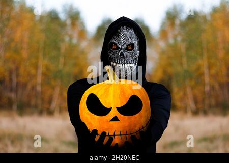 Grim reaper tenant la tête de citrouille d'halloween.Homme dans le masque de la mort avec une flamme de feu dans les yeux sur fond de forêt d'automne jaune.Halloween concept de vacances.Dar Banque D'Images