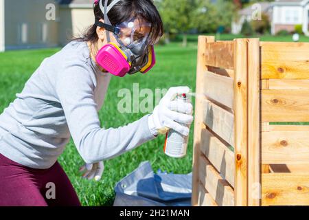 Une jeune femme caucasienne portant des lunettes de sécurité et des gants masque est en spray tachant une clôture en bois à l'extérieur.Image de concept pour projet diy et HEAL Banque D'Images
