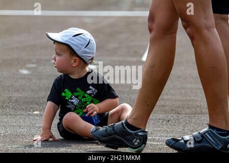 Columbus, OH États-Unis 08-07-2021: Un jeune garçon blond fatigué assis sur le sol dans un parking pendant un événement que ses parents sont vus à côté de lui.Il Banque D'Images