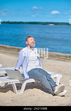 Homme se détendant près d'un ordinateur portable sur une chaise longue de plage Banque D'Images