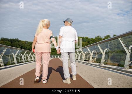 Une femme et un homme âgés positifs marchent en tenant les mains le long de la passerelle après l'entraînement Banque D'Images