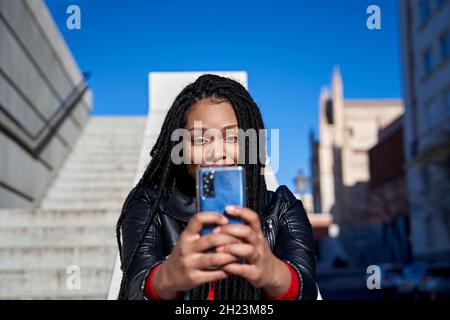 Portrait d'une fille afro-américaine avec des dreadlocks dans ses cheveux envoyant des messages avec son téléphone cellulaire.Concept de technologie Banque D'Images