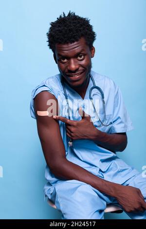 Une infirmière afro-américaine qui pointe vers le bandage de vaccins contre les vaccins tout en regardant la caméra en studio.Assistant médical noir montrant un pansement adhésif après avoir été vacciné contre le coronavirus Banque D'Images