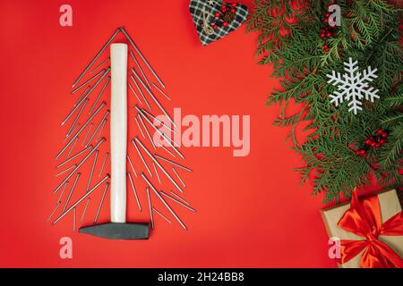 Clous longs et marteau, disposés en forme d'arbre de Noël avec branches d'épinette et boîte cadeau sur fond rouge.Pose à plat Banque D'Images