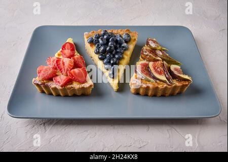 Gros plan sur trois desserts de gâteau à tarte avec fraises, bleuets et figues sur une assiette rectangulaire sur fond texturé clair Banque D'Images