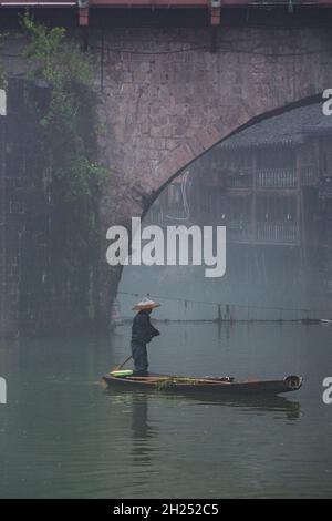 Un homme pagaie un sampan dans le brouillard près du Phoenix Hong Bridge sur la rivière Tuojiang, Fenghuang, Chine. Banque D'Images