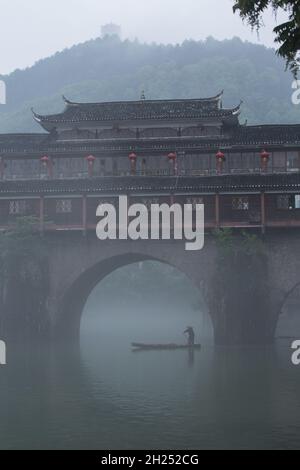 Un homme pagaie un sampan dans le brouillard près du Phoenix Hong Bridge sur la rivière Tuojiang, Fenghuang, Chine. Banque D'Images