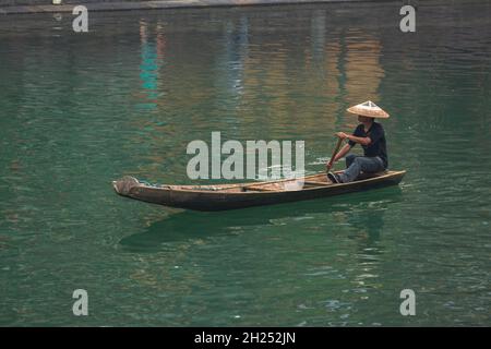 Un homme dans un chapeau conique traditionnel pagaie un sampan sur la rivière Tuojiang à Fenghuang, en Chine. Banque D'Images