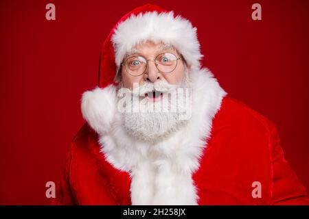 Photo de drôle impressionné homme à la retraite porter santa claus costume lunettes à bouche ouverte célébrant noël isolé couleur rouge fond Banque D'Images