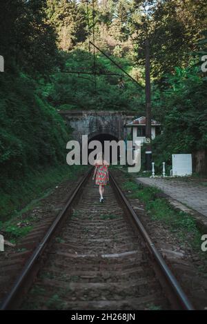 Une jeune femme en robe colorée marche le long des rails dans la forêt.Une fille solitaire s'approche d'un tunnel dans la forêt verte.Vue arrière. Banque D'Images