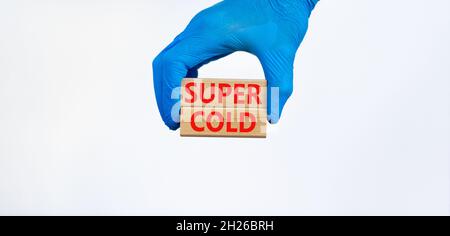 Symbole Super froid.Mots Super froid sur des blocs de bois.Main médecin dans un gant bleu.Magnifique fond blanc.Concept médical, Covid-19 et super froid. Banque D'Images