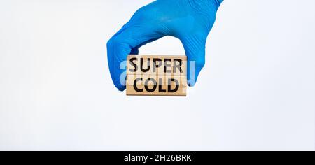 Symbole Super froid.Mots Super froid sur des blocs de bois.Main médecin dans un gant bleu.Magnifique fond blanc.Concept médical, Covid-19 et super froid. Banque D'Images