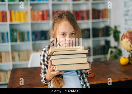 Portrait de taille moyenne d'une adorable fille de l'école primaire tenant une pile de livres dans la bibliothèque à l'école regardant un appareil photo. Banque D'Images