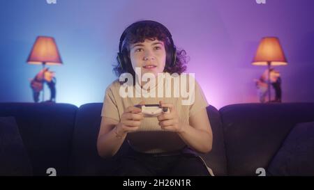 Portrait d'une jeune femme de joueur attirante assise sur un canapé, jouant à un jeu vidéo en ligne avec une manette sans fil sur la console à la maison.Jeune gamer fille W Banque D'Images