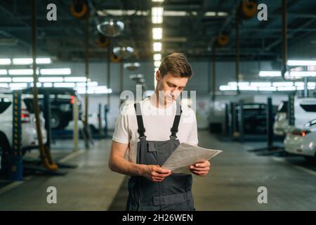 Vue avant de la belle et ciblée jeune mécanicien homme portant un écritoire uniforme de lecture debout dans le garage de l'atelier de réparation automobile Banque D'Images