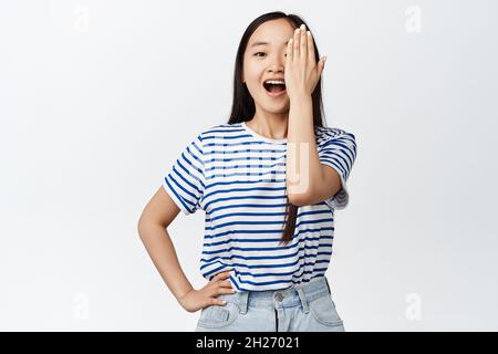 Concept de style de vie et d'émotions.Happy asian girl couvre une moitié de visage, regardant l'appareil photo surpris, bouche ouverte dans la crainte, se tenant étonné contre Banque D'Images