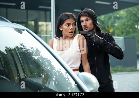 Voleur en colère menaçant avec le pistolet de peur fille.L'homme européen et la jeune femme brunette se tiennent près de l'automobile.Veste à capuche bandit pour homme.Concept Banque D'Images