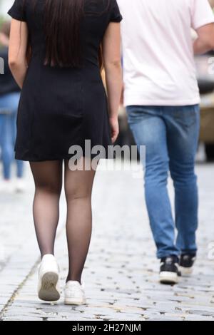 Junge Frau mit schwarzem Minikleid und schwarzen Strünpfen geht hinter einem Mann in Jeans und weißem T-shirt - Jeune femme avec mini robe noire et Banque D'Images