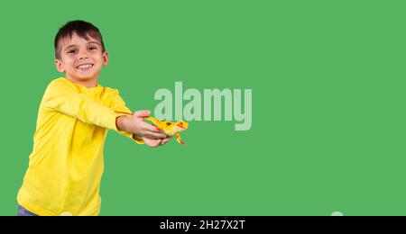 Un petit garçon malin à capuchon jaune avec un sourire sur son visage prêt sa main avec un jouet de dinosaure, debout contre un studio de fond vert, espace de copie. Banque D'Images