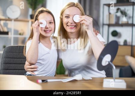 Une mère et une fille caucasiennes souriant sur l'appareil photo tout en cachant un œil derrière un tampon de coton.Femme et adolescent appréciant des rituels de beauté à la maison. Banque D'Images