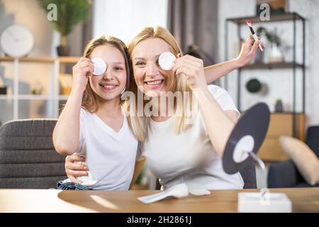 Une mère et une fille caucasiennes souriant sur l'appareil photo tout en cachant un œil derrière un tampon de coton.Femme et adolescent appréciant des rituels de beauté à la maison. Banque D'Images