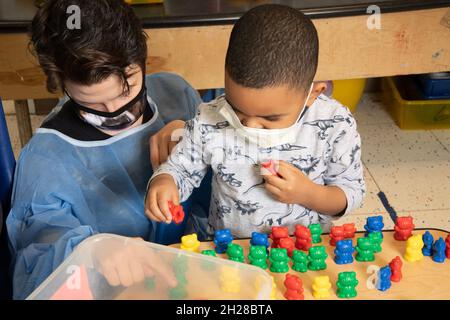 Éducation préscolaire 4-5 ans garçon triant et alignant les ours en plastique coloré par couleur, jeune enseignant regardant sur, les deux portant des masques de visage Covid-19 Banque D'Images