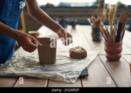 Gros plan d'un artisan méconnaissable debout sur un bureau en bois avec du polyéthylène et de l'argile à couper avec coupe-fil dans un atelier de poterie Banque D'Images