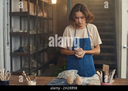 potier jeune femelle concentré dans le tablier en train de baller l'argile tout en le préparant pour sculpter dans l'atelier Banque D'Images