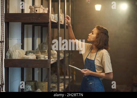 Jeune caucasien sérieux potter dans le tablier de denim tenant le comprimé et examinant les pots d'argile sur la tablette dans l'atelier sombre Banque D'Images